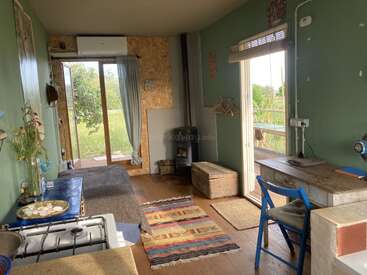 This cozy tiny house features green walls, a wood stove, rustic furniture, colorful rugs, a compact kitchen, natural light, and views of lush greenery outside.