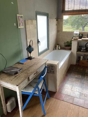 A rustic bathroom features a bathtub by a window, a small wooden desk with a blue chair, houseplants, mosaic tiles, and cozy, eclectic decor.