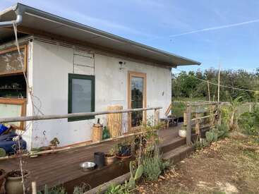 A small house with a wooden porch, surrounded by plants and garden. The exterior is simple, rustic, and set against a clear blue sky.