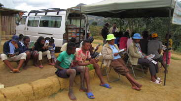 La imagen muestra a un grupo de personas reunidas en torno a una mesa, con algunos individuos sentados y otros de pie, todos mirando en la misma dirección, en una habitación con una gran ventana.