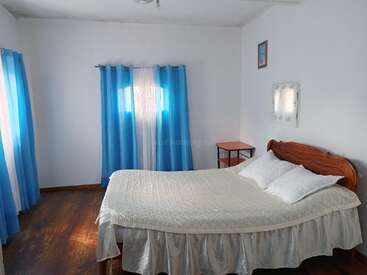 La imagen muestra un dormitorio sereno con paredes blancas, suelos de madera oscura y una cama adornada con un edredón blanco, dos almohadas y un cabecero de madera.