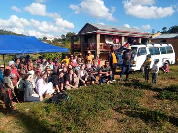 La imagen muestra a un grupo de personas reunidas frente a una casa, con una furgoneta blanca y una tienda de campaña azul cerca, sobre un fondo de cielo azul brillante con nubes esponjosas.