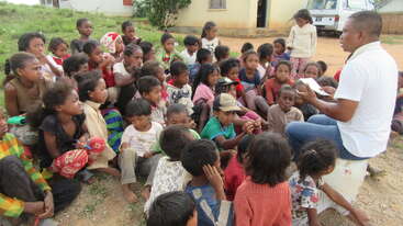 Un grupo de niños se reúnen en torno a un hombre, probablemente un profesor o líder, formando un círculo. La escena parece ser un aula o un entorno educativo. Todos los niños miran hacia el hombre.
