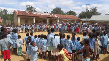 La imagen muestra a un grupo de niños reunidos en círculo, probablemente participando en una actividad escolar, con algunos adultos presentes, lo que transmite una sensación de comunidad y aprendizaje.