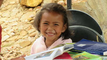 La imagen muestra a una joven sentada en el suelo, rodeada de libros y papeles, con un fondo neutro. El ambiente general es de sencillez y minimalismo.