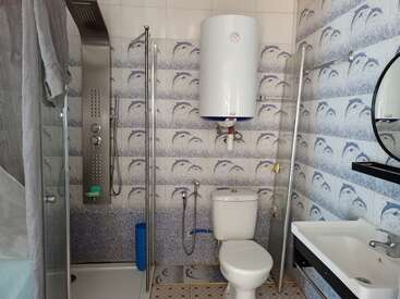 La imagen muestra un cuarto de baño compacto con ducha, inodoro y lavabo, con una pared de azulejos con motivos de delfines y un calentador de agua blanco montado en la pared.