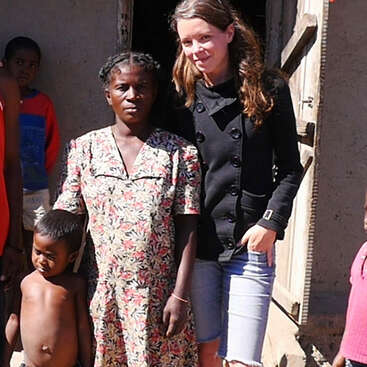 La imagen muestra a una mujer con chaqueta negra junto a un niño y una mujer mayor delante de un edificio, rodeados de otras personas.