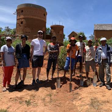 La imagen muestra a un grupo de personas de pie en un campo de tierra, posando para una foto con palas, delante de tres estructuras de ladrillo y un cielo azul.