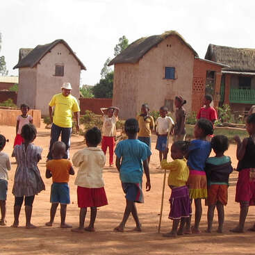La imagen muestra a un grupo de niños jugando en un campo, con algunos adultos presentes, probablemente durante un acto escolar o una reunión comunitaria, transmitiendo una sensación de alegría y comunidad.