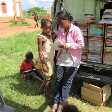 Das Bild zeigt eine Gruppe von Menschen, die um einen Tisch versammelt sind. Einige Personen halten Bücher oder Papiere in der Hand, während andere auf etwas auf dem Boden schauen. Die Szene scheint ein Treffen oder eine Diskussion zu sein.