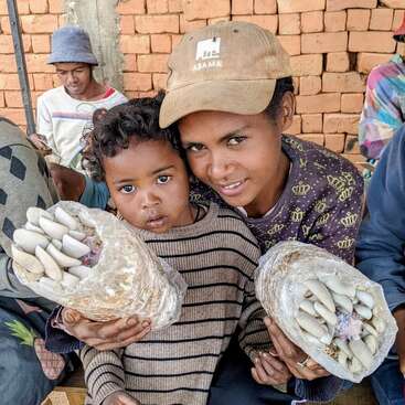 Una mujer y un niño sonríen, sentados muy juntos, cada uno con una bolsa de setas en la mano. Otras personas están sentadas cerca, con una pared de ladrillos al fondo, vestidas con ropa informal. Ambiente cálido.