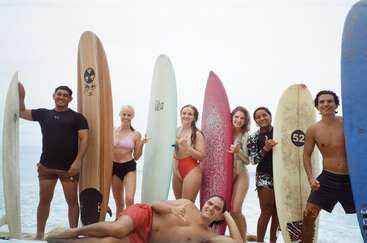 A group of seven people stand with surfboards on a beach, smiling and posing. One person lies in front, everyone looks happy, ready to surf.