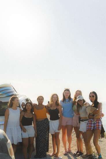 Eight young women stand together by a car with surfboards, smiling on a sunny beach day. One woman holds a small dog, the sky is bright.