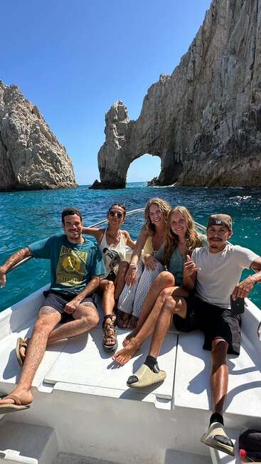 Five friends are sitting on a boat, smiling and enjoying sunny weather. Behind them are dramatic rocky cliffs and the famous Arch of Cabo San Lucas.