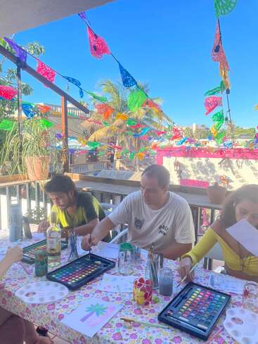 Three people are painting at a colorful outdoor table covered with art supplies. Festive papel picado banners hang above, and palm trees are in the background.
