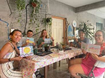 Five people sit around a decorated table, happily showing their colorful artwork. Art supplies are scattered on the table. Behind them, plants and a wall map decorate the room.