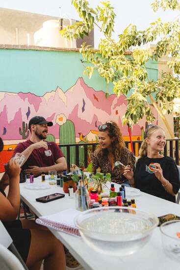 Four people sit around a table outdoors, laughing and painting. Bright paint bottles and brushes fill the table. A colorful, desert-themed mural decorates the background wall.