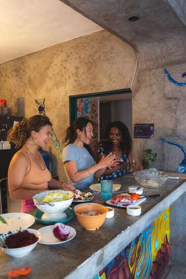 Three women are happily chatting and preparing food in a cozy, colorful kitchen. Fresh ingredients and bowls are spread across the counter, creating a lively, friendly atmosphere.