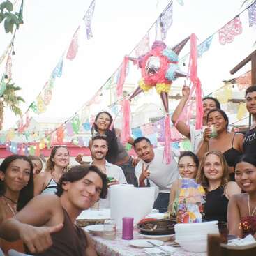 Un alegre grupo de amigos celebra al aire libre bajo coloridas pancartas de papel picado y una piñata, compartiendo comida y sonrisas en una reunión festiva y animada a la luz del día.