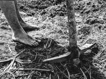 L'image représente une personne pieds nus, debout sur le sol, à côté d'un grand outil métallique rouillé, doté d'un long manche et d'une lame tranchante, entouré de terre et de débris.