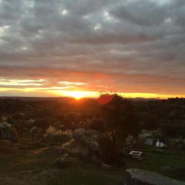 L'image représente un coucher de soleil serein sur un paysage rural, avec un ciel nuageux, des arbres et des rochers, ainsi qu'une petite structure blanche visible au premier plan.