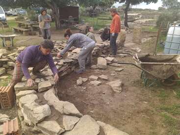 L'image représente un groupe de personnes construisant un mur de pierre en plein air, avec une brouette et d'autres outils à proximité.