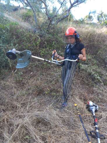 Une femme portant un casque et un équipement de sécurité se tient dans un champ, tenant un coupe-bordure muni d'une longue perche et d'une grande tête de coupe.