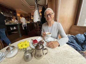 Una anciana con gafas disfruta de un café y un pastel en una acogedora mesa de cafetería, adornada con flores amarillas, nata y azúcar. La cafetería bulle de actividad detrás de ella.