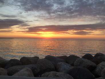 Una serena puesta de sol sobre aguas tranquilas, con grandes rocas en primer plano. El cielo está pintado con tonos anaranjados, rosas y morados, que crean una atmósfera apacible.