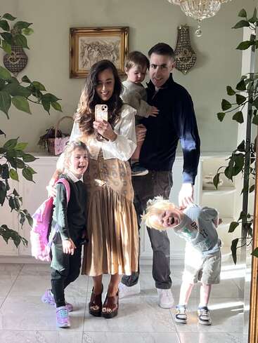 Una familia de cinco miembros posa en una habitación luminosa, sonriendo en un selfie en el espejo. Los padres sostienen a dos niños; delante, una niña con mochila.