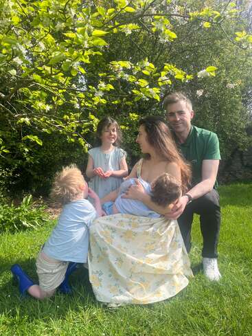 Una familia de cinco miembros está sentada junta sobre la hierba verde bajo un árbol en flor. Sonríen e interactúan, disfrutando de un día soleado en la naturaleza.