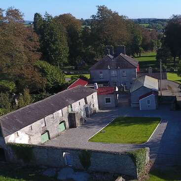 The image depicts a large estate with a sprawling lawn and a long driveway, featuring a substantial house with multiple gables and a prominent roof. The property is surrounded by a fence.