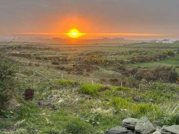 A stunning sunrise casts warm orange light over a misty rural landscape with grassy fields, scattered bushes, distant buildings, and a rustic stone wall in front.