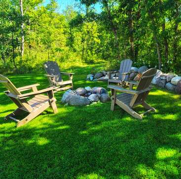 Quatre chaises Adirondack entourent un foyer en pierre sur une pelouse verdoyante, au milieu d'une clairière ensoleillée et paisible, avec des arbres et des rochers empilés en arrière-plan.
