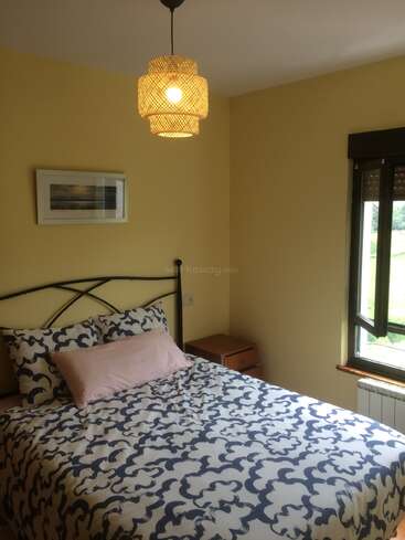 Une chambre à coucher confortable avec des murs crème, une literie à motifs, un oreiller rose pâle, une œuvre d'art encadrée, une table de nuit en bois, un luminaire tressé et une fenêtre ouverte.