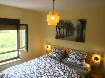 Une chambre confortable avec un lit double, une literie à motifs, une tête de lit en bois avec des lumières intégrées, une fenêtre ouverte, des murs jaunes, une vue sur la nature et une œuvre d'art représentant un arbre serein.