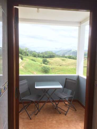 Un balcon confortable avec deux chaises pliantes grises et une petite table en verre donne sur un paysage verdoyant et des collines lointaines sous un ciel nuageux. Ambiance paisible.