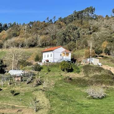 Ein weißes Haus mit rotem Dach steht auf einem Hügel, umgeben von grünem Gras, Bäumen, einem Garten, blauem Himmel und einem bewaldeten Gebiet in der Ferne. Eine friedliche ländliche Szenerie.
