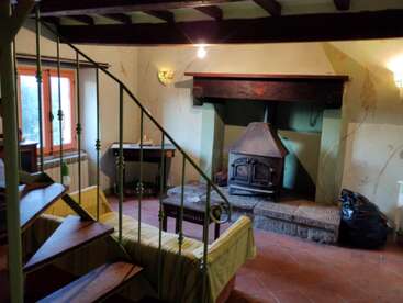 A cozy rustic living room with a wood-burning fireplace, spiral staircase, exposed wooden beams, yellow sofa, large window, tiled floor, and soft, warm lighting.