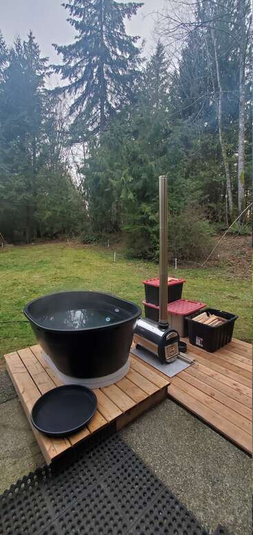 A black outdoor hot tub sits on a wooden deck, surrounded by trees. Nearby, a wood-burning stove and firewood bins add rustic charm and warmth.