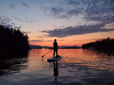 A person paddleboards across calm water during a stunning sunset, surrounded by silhouetted trees and distant mountains, reflecting beautiful orange, pink, and purple skies. Peaceful tranquility.