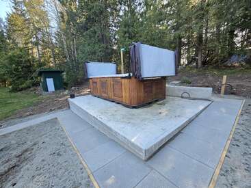 This image shows a wooden hot tub with its cover partially open, sitting on a concrete pad, surrounded by trees and nature in a peaceful setting.