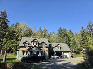 A charming two-story house with dormer windows sits amidst tall green trees. Two cars are parked outside under a clear blue sky, suggesting a peaceful setting.
