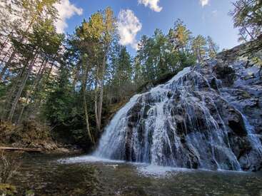 A stunning waterfall cascades down rocks surrounded by tall green trees. Sunlight filters through the branches, with a clear blue sky and fluffy clouds above.