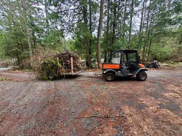 A utility vehicle is towing a trailer loaded with tree branches and yard debris through a forested area on a dirt and gravel road, surrounded by trees.