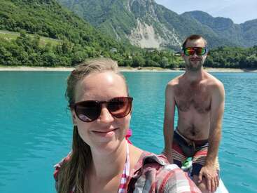 L'image représente une scène sereine d'un homme et d'une femme debout dans une étendue d'eau, avec un paysage montagneux en arrière-plan et des arbres le long de la rive.
