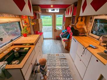 L'intérieur chaleureux et coloré d'un camping-car met en scène un bébé qui explore les tiroirs de la cuisine, un chat sur le plan de travail, une guitare, une vue sur des champs verdoyants depuis la fenêtre et une décoration ludique.