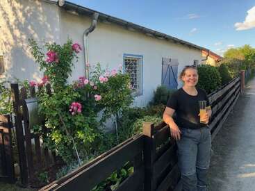 A smiling woman stands by a wooden fence, holding a drink. Pink flowers and greenery surround a quaint house with blue window frames, under clear skies.