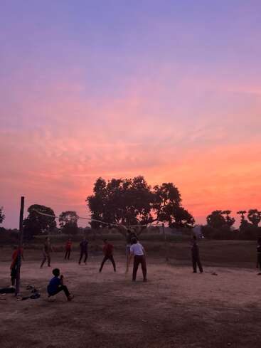 Un grupo de personas juega al voleibol en una pista polvorienta al aire libre durante la puesta de sol, con cielos de vibrantes colores rosa y morado y siluetas de árboles al fondo.