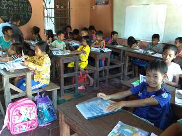 The image depicts a classroom with approximately 20 children seated at wooden desks, engaged in learning activities, surrounded by books and backpacks, with a teacher present in the background.